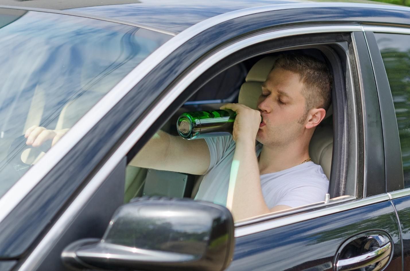 A young man sits behind the steering wheel of a dark-colored car, drinking alcohol from a green glass bottle.