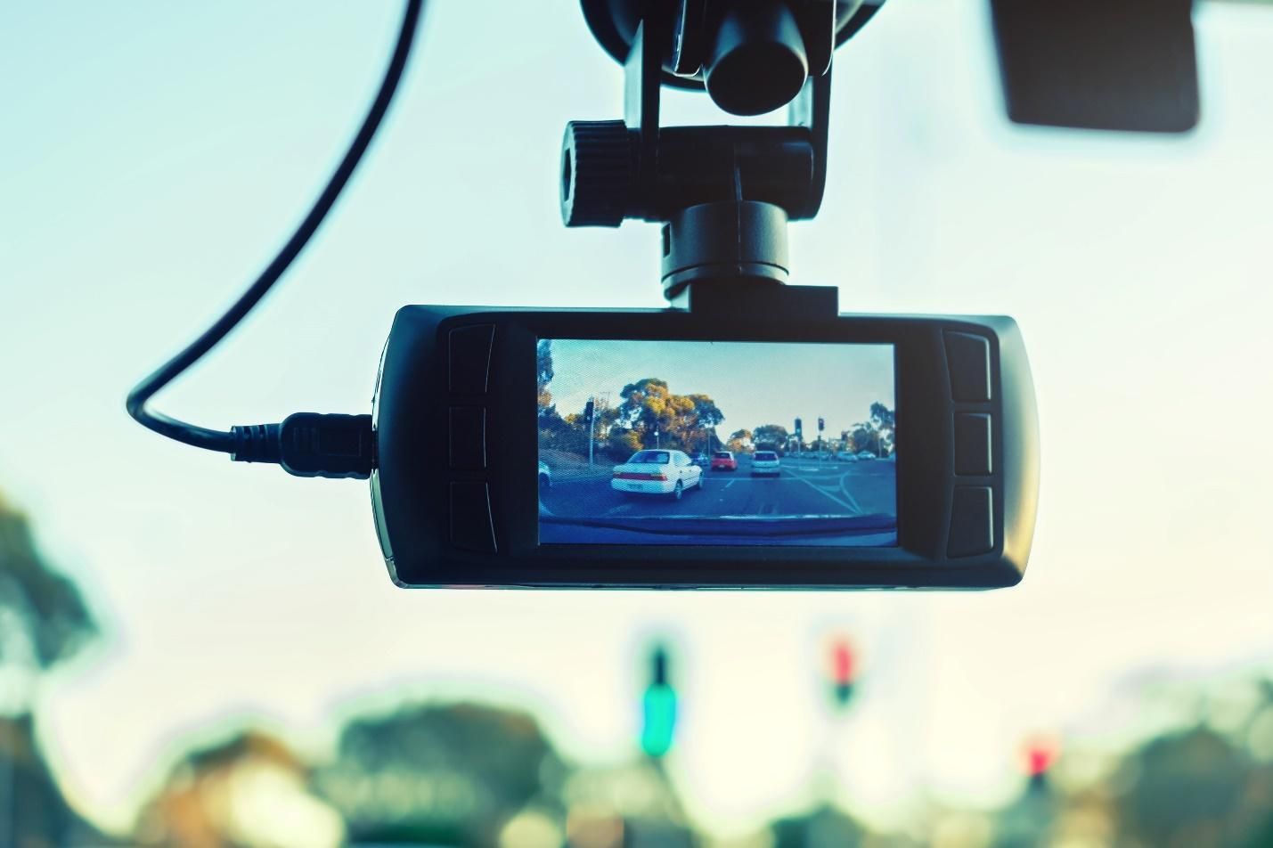 A small rectangular camera with a power cord plugged into one side, hangs from a suction cup on a car’s windshield.