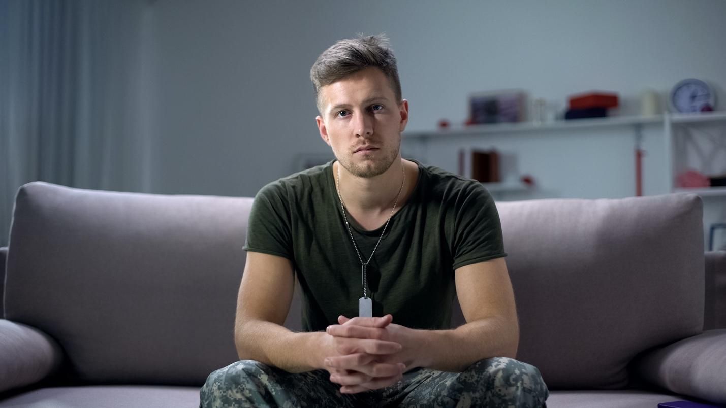 Military serviceman in fatigues, sitting on a sofa with hands clasped.