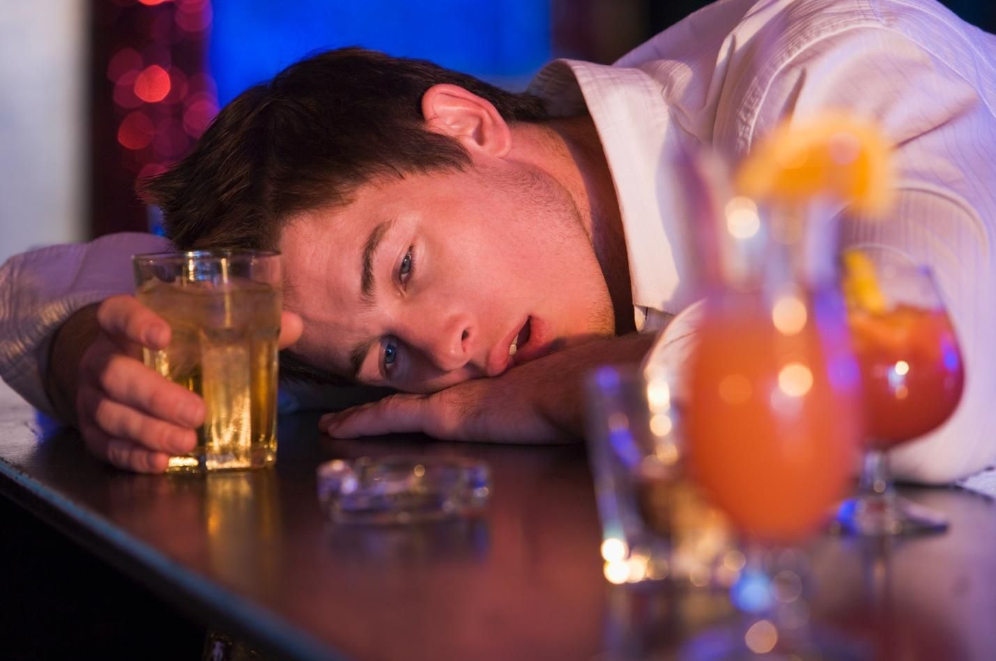 An intoxicated young man in a collared white button-down business shirt rests his head on a bar next to his drink; high blood alcohol concentration concept.