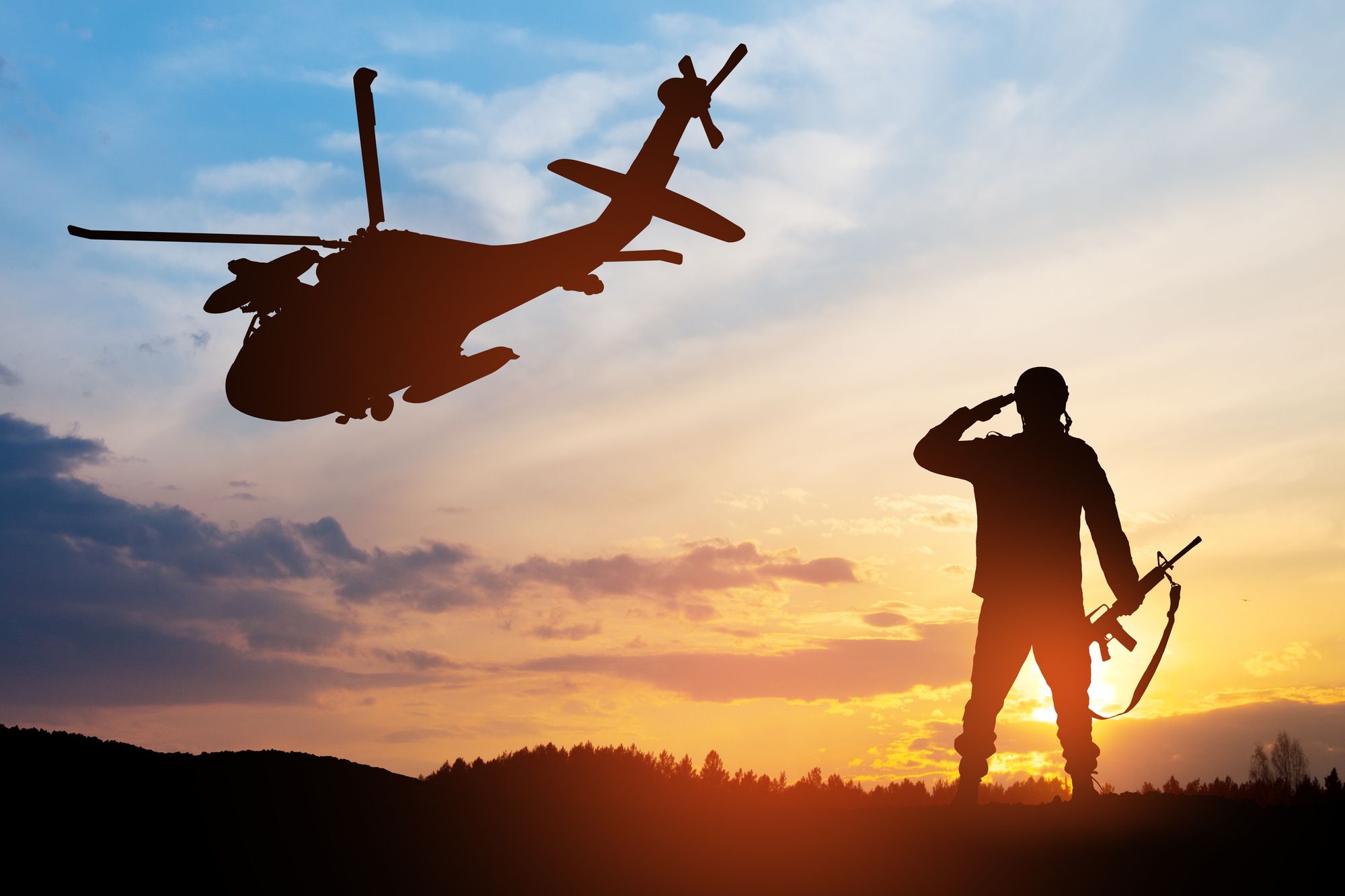 Silhouette view of a soldier on military deployment saluting a departing helicopter at sunset.