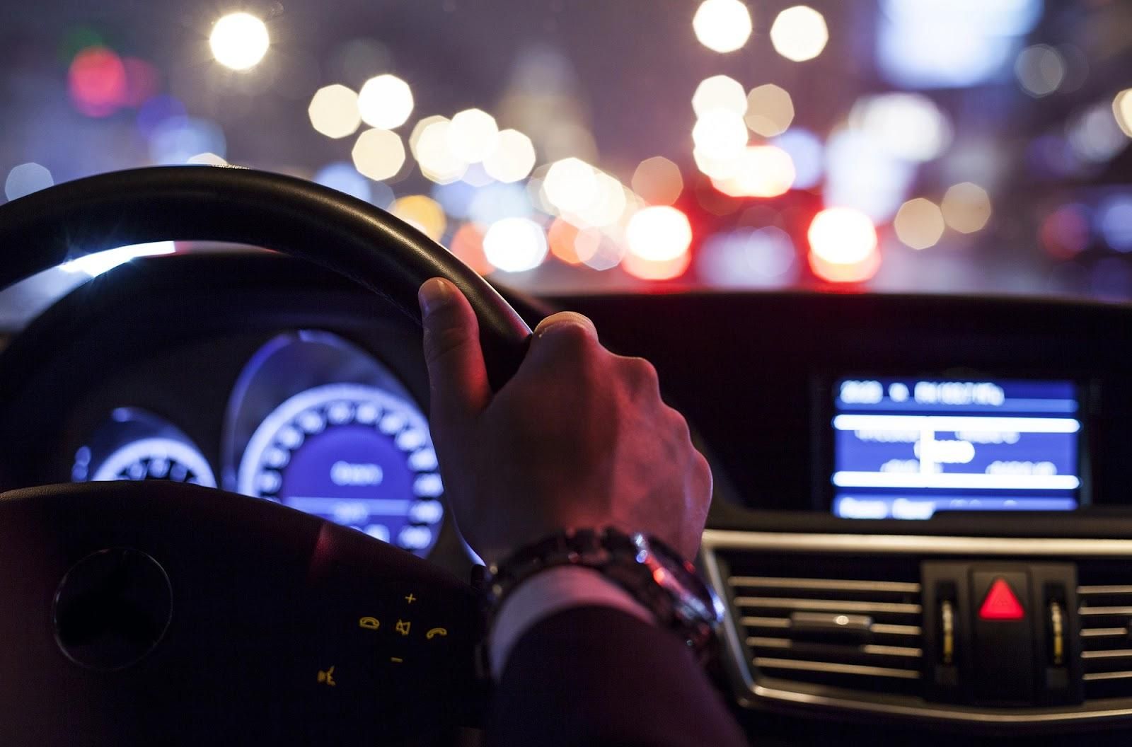 A man driving with city nightlife outside the windshield.