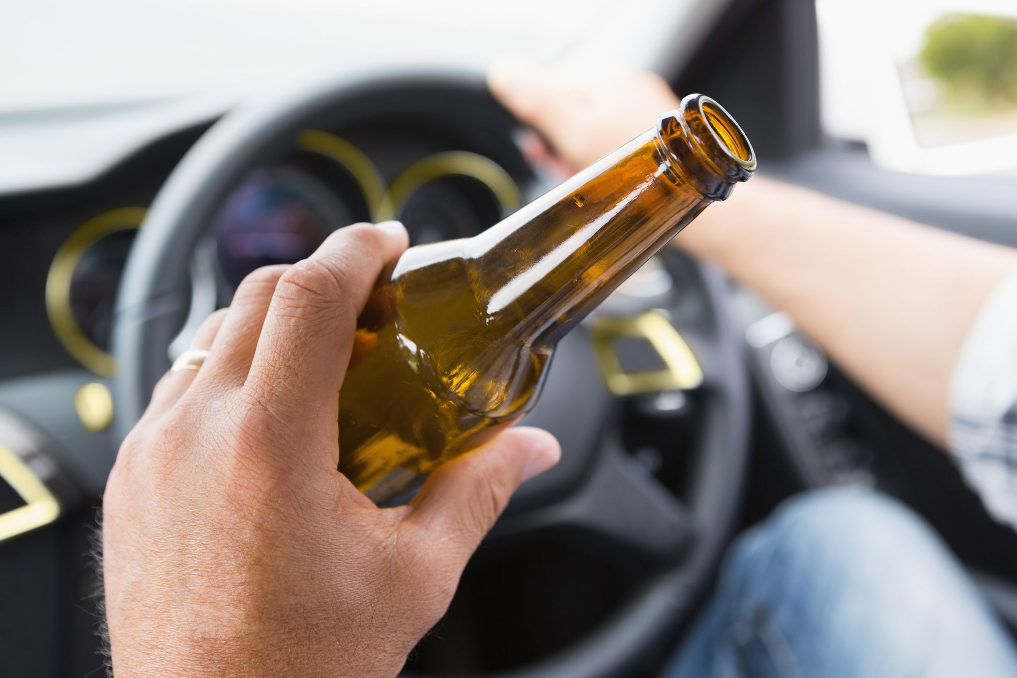 Closeup of a man’s hand holding a beer bottle in the foreground; background shows blurred image of steering wheel.