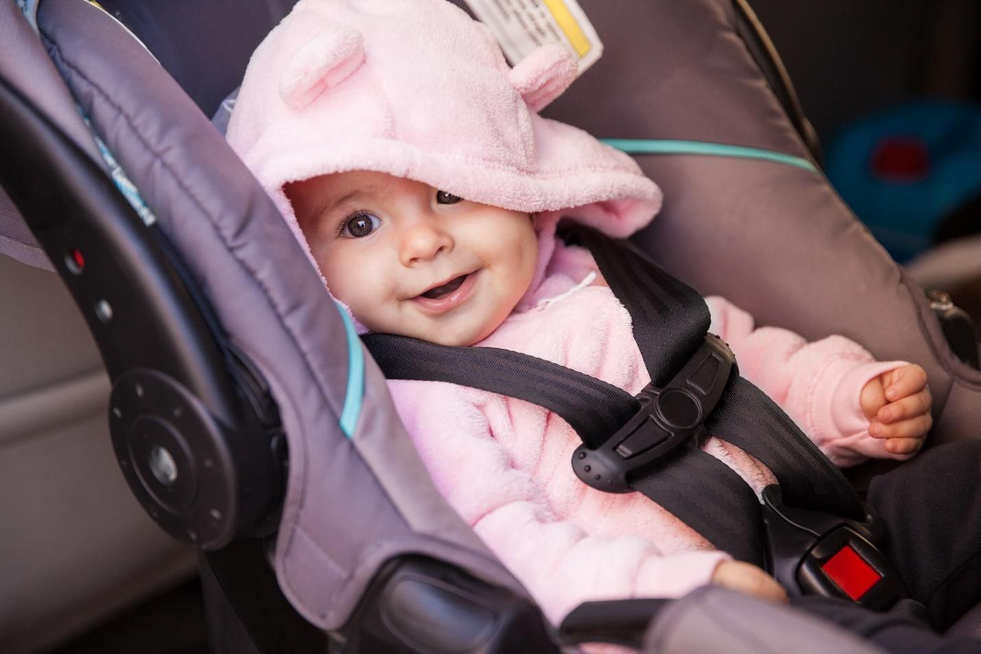 A baby dressed in pink smiling and strapped into a car seat.