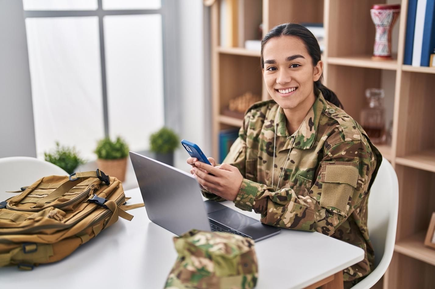 Smiling young woman in military fatigues typing on smartphone in front of laptop computer.